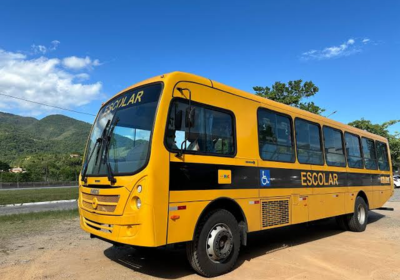 Ônibus escolar amarelo e preto estacionado em estrada de terra, com acessibilidade indicada por símbolo na lateral. Ao fundo, vegetação verde, montanhas e céu azul com nuvens brancas.