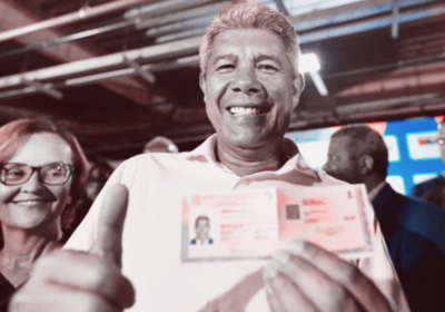 Governador da Bahia, Jerônimo Rodrigues, sorri e exibe uma carteira de identidade em evento oficial de lançamento do novo RG nacional, enquanto outras autoridades aparecem ao fundo. A imagem simboliza a campanha de modernização dos documentos, apesar da escassez de material para emissão do RG em cidades do interior baiano.