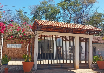 Fachada da sede da APAE de Correntina, com portão de grades pretas, cobertura de telhas e jardim com plantas e flores, em um dia ensolarado.